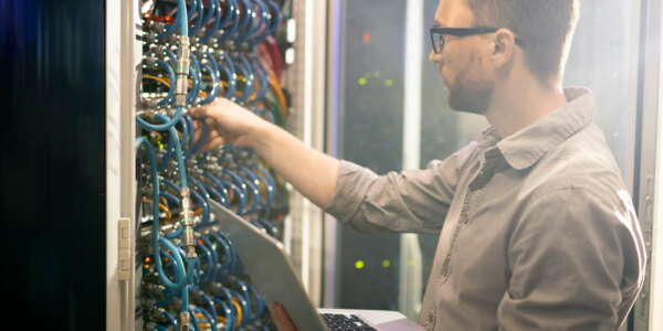 Serious busy young male engineer in glasses standing against mainframe and adjusting cables while analyzing server connections